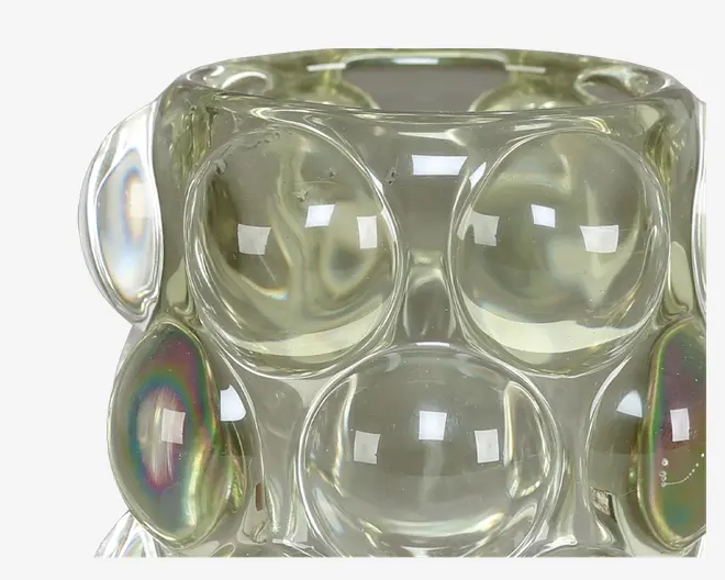 Nærbillede af en gennemsigtig glasgenstand med en tekstureret overflade, der har store, bulede boble-lignende forhøjninger. Glasset har et let gulligt skær og reflekterer lyset, hvilket skaber iriserende højdepunkter og regnbueeffekter på nogle af boblerne. Overfladen er glat og blank.