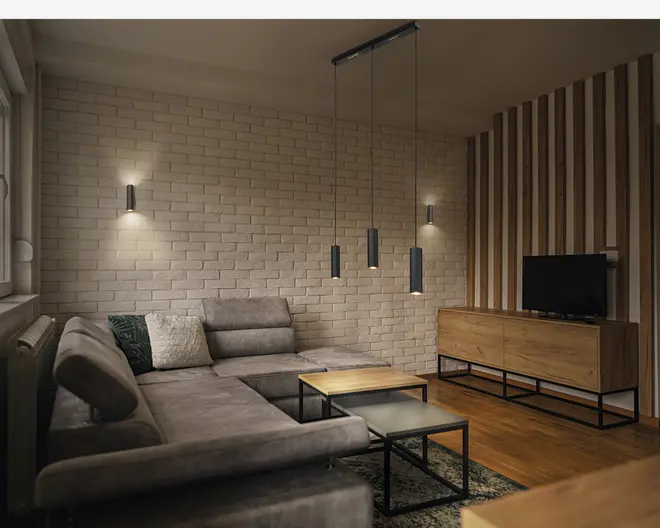 Moderne stue med en stor L-formet grå hjørnesofa med bløde puder, et sæt af tre indskudsborde med sorte metalstel og trætoppe samt et lavt tv-møbel i træ med sorte metalben. Rummet har en hvidmalet murstensvæg, lodrette træpaneler på den ene side og moderne pendellamper hængende fra loftet. Der er også to vægmonterede væglamper, der giver blødt, stemningsfuldt lys. Gulvet er i træ, og et mønstret tæppe er delvist synligt under sofabordene.