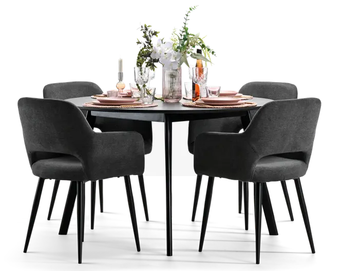 Rundt sort spisebord med fire mørkegrå polstrede lænestole, dækket op med rosa tallerkener, glas og et højt blomsterarrangement i hvidt og grønt.