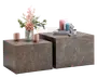Sæt med to firkantede sideborde i forskellige højder med marmorlignende overflade i brun med diskrete hvide årer. Bordene har et moderne, minimalistisk kube-design. Det ene bord er pyntet med en glasvase med lyserøde og hvide blomster samt to lyserøde kopper, mens det andet har en stak bøger, dekorative lys i lyserød og blå samt en lille abstrakt skulptur.