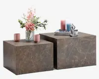 Sæt med to firkantede sideborde i forskellige højder med marmorlignende overflade i brun med diskrete hvide årer. Bordene har et moderne, minimalistisk kube-design. Det ene bord er pyntet med en glasvase med lyserøde og hvide blomster samt to lyserøde kopper, mens det andet har en stak bøger, dekorative lys i lyserød og blå samt en lille abstrakt skulptur.