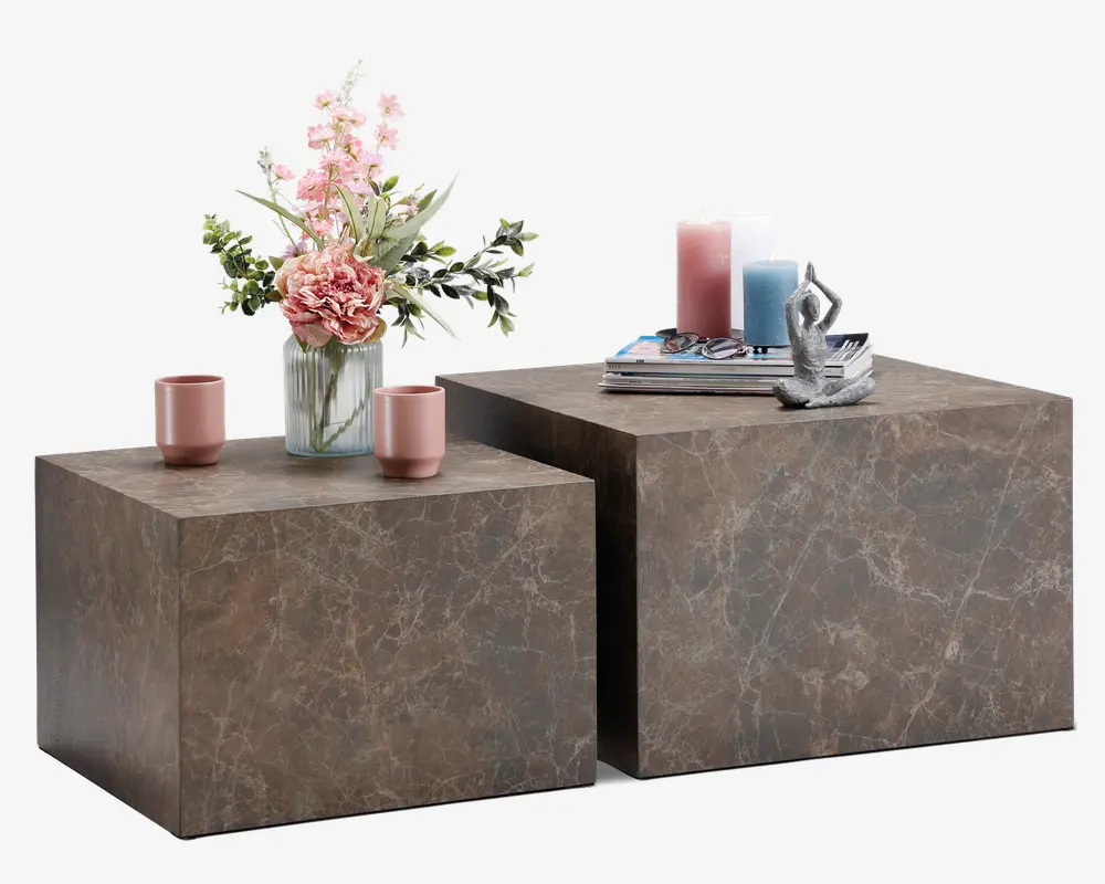 Sæt med to firkantede sideborde i forskellige højder med marmorlignende overflade i brun med diskrete hvide årer. Bordene har et moderne, minimalistisk kube-design. Det ene bord er pyntet med en glasvase med lyserøde og hvide blomster samt to lyserøde kopper, mens det andet har en stak bøger, dekorative lys i lyserød og blå samt en lille abstrakt skulptur.
