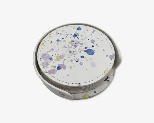 Rund genstand med en hvid overflade, der har et farverigt stænkmønster i blå, gul og lilla. Genstanden har synlige syninger langs kanten og opbevares i en matchende holder. Designet er moderne og kunstnerisk med en glat finish og en kompakt, stabelbar form.