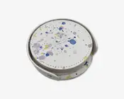 Rund genstand med en hvid overflade, der har et farverigt stænkmønster i blå, gul og lilla. Genstanden har synlige syninger langs kanten og opbevares i en matchende holder. Designet er moderne og kunstnerisk med en glat finish og en kompakt, stabelbar form.