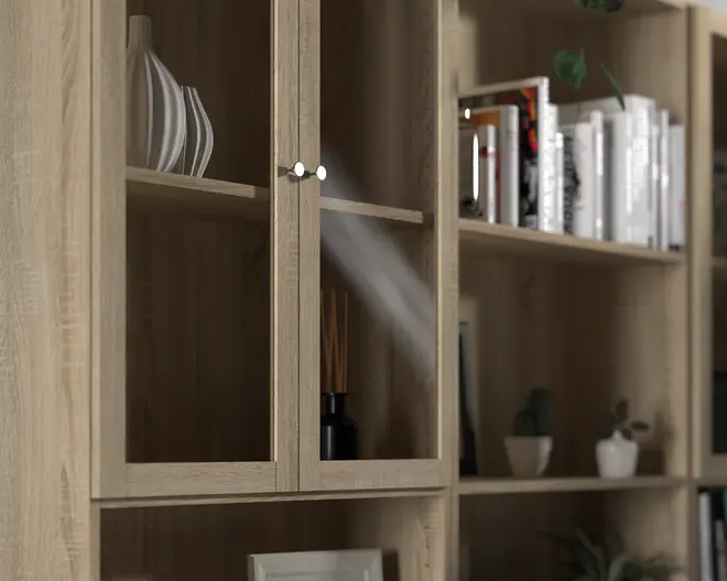 Nærbillede af en reol i lyst egetræslook med en sektion med glaslåger og runde sølvfarvede greb. Hylderne er dekoreret med hvide keramikvaser i moderne, bølget design, en sort duftspreder med pinde, bøger med hvide og sorte rygge samt små potteplanter. Træets årestruktur er synlig, og stilen er moderne og minimalistisk.