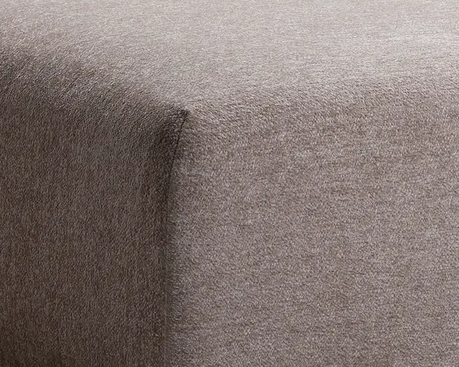 Nærbillede af hjørnet på et møbel betrukket med blødt, taupefarvet stof. Stoffet har en glat, fløjlsagtig tekstur og er stramt tilpasset rundt om kanterne, hvilket giver et rent og moderne udtryk.
