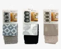 Tre indpakkede badesæt, hver med et badeforhæng, bademåtte og gardinringe. Det første sæt har et geometrisk diamantmønster i grå nuancer med en matchende lysegrå tekstureret måtte. Det andet sæt har et moderne sort-hvidt stribet forhæng med en ensfarvet sort tekstureret måtte. Det tredje sæt viser et taupefarvet forhæng med diskrete vanddråbe-designs og en beige tekstureret måtte. Hvert sæt indeholder tre gardinringe i matchende farver.