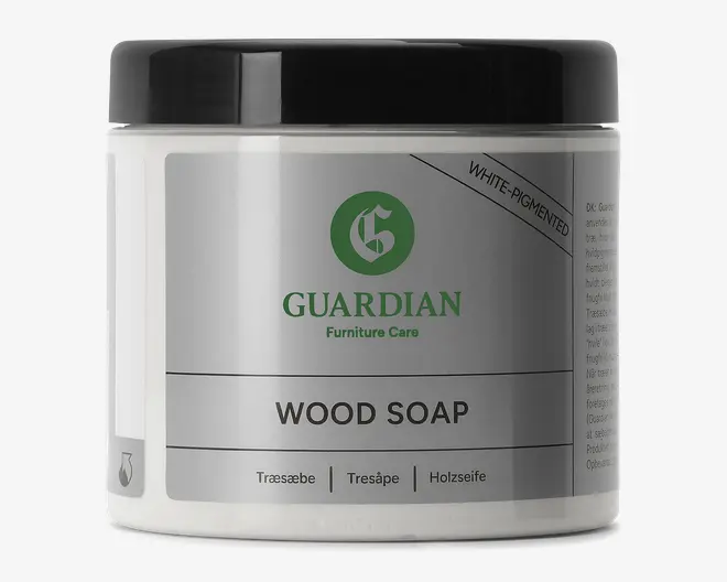 En rund plastbeholder med sort skruelåg og sølvfarvet/hvid etiket. Etiketten viser mærket 'Guardian Furniture Care' i grøn og angiver, at produktet er 'Wood Soap' med hvid pigmentering. Der er også oversættelser af 'træsæbe' på dansk, norsk og tysk. Beholderen ser robust ud og egnet til opbevaring af rengøringsprodukter.