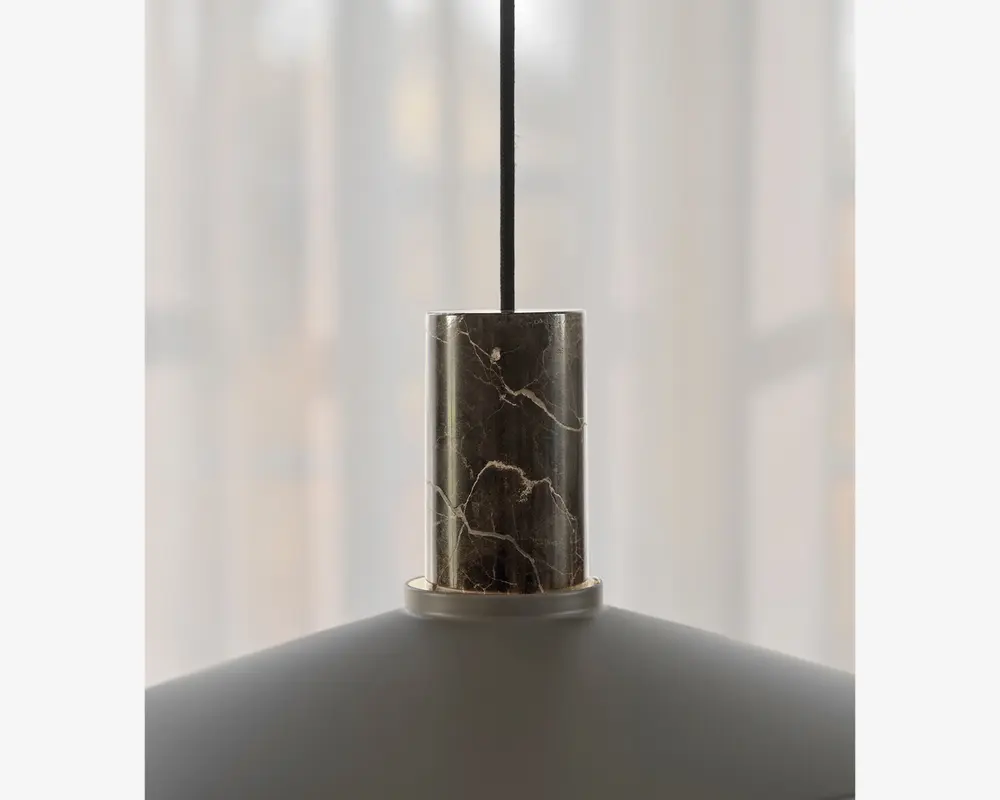Nærbillede af den øverste del af en pendellampe, der har en cylindrisk marmordetalje med naturlige hvide årer. Marmorelementet forbinder den sorte ophængssnor med den matte lampeskærm nedenunder, hvilket giver lampen et moderne og elegant udtryk.