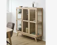 Et vitrineskab i lyst træ med fire glaslåger og glaspaneler på siderne, udstyret med tre indvendige glashylder til udstilling. Skabet står på skråtstillede ben og har minimalistiske, lodrette greb. Indeni er der arrangeret forskellige tallerkener, skåle og dekorative genstande. På toppen står en lampe, en lille skål og et par bøger, hvilket giver et moderne udtryk.