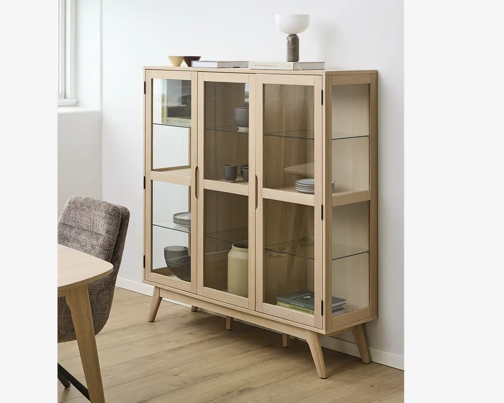 Et vitrineskab i lyst træ med fire glaslåger og glaspaneler på siderne, udstyret med tre indvendige glashylder til udstilling. Skabet står på skråtstillede ben og har minimalistiske, lodrette greb. Indeni er der arrangeret forskellige tallerkener, skåle og dekorative genstande. På toppen står en lampe, en lille skål og et par bøger, hvilket giver et moderne udtryk.