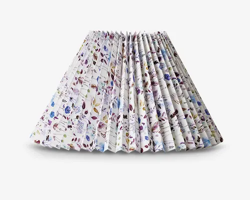Plisseret lampeskærm med bred, konisk form. Stoffet har et fint blomstermønster i nuancer af lilla, blå og grøn på hvid baggrund. Plisseringerne giver et tekstureret og struktureret udtryk, og blomstermotivet er jævnt fordelt over hele overfladen.