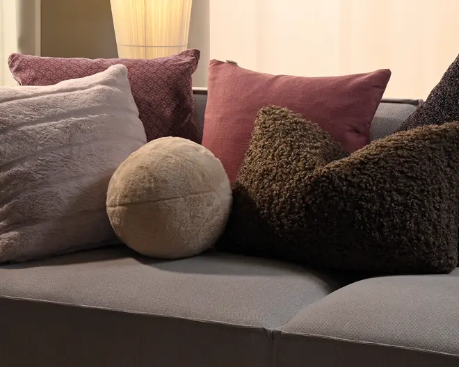 En grå sofa stylet med flere pyntepuder: en stor lysegrå blød pude, en mønstret mauve pude, en støvet rosa rektangulær pude, en lille rund beige pude samt to mørkebrune teksturerede puder med et kruset, bouclé-lignende udtryk.
