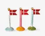 Tre dekorative flagstænger i keramik, hver med et gyldent hjerte på toppen og et lille dansk flag. Flagstængerne har forskellige farvede fod: lyseblå, orange og olivengrøn. Ordet 'TILLYKKE' står skrevet lodret på hver stang. Designet er festligt og velegnet til fejringer.