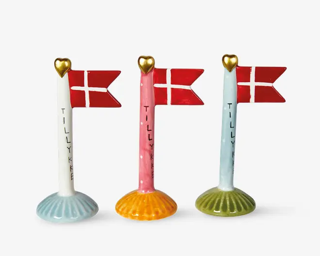 Tre dekorative flagstænger i keramik, hver med et gyldent hjerte på toppen og et lille dansk flag. Flagstængerne har forskellige farvede fod: lyseblå, orange og olivengrøn. Ordet 'TILLYKKE' står skrevet lodret på hver stang. Designet er festligt og velegnet til fejringer.
