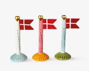 Tre dekorative flagstænger i keramik, hver med et gyldent hjerte på toppen og et lille dansk flag. Flagstængerne har forskellige farvede fod: lyseblå, orange og olivengrøn. Ordet 'TILLYKKE' står skrevet lodret på hver stang. Designet er festligt og velegnet til fejringer.