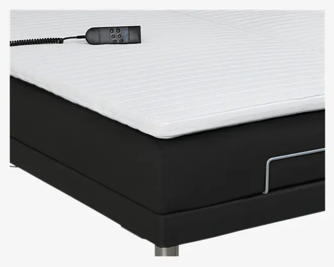Nærbillede af et sengehjørne med en tyk, hvid, quiltet topmadras med horisontale syninger, der ligger på en sort polstret bund. En ledningsforbundet fjernbetjening med flere knapper ligger på sengen, hvilket indikerer justerbare funktioner. Sengen har en metalstang på siden og står på cylindriske metalben.