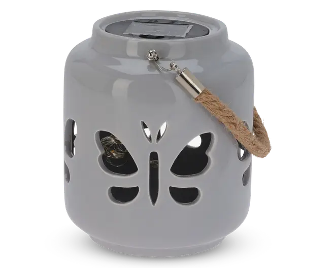 En rund, blank keramiklanterne med udskårne sommerfugleformer på siderne. Den har et robust rebhåndtag fastgjort med metalbeslag og et solpanel på toppen til opladning. Indvendigt ses en lille pære gennem udskæringerne, som er designet til at skabe dekorative lysmønstre, når lanternen er tændt.