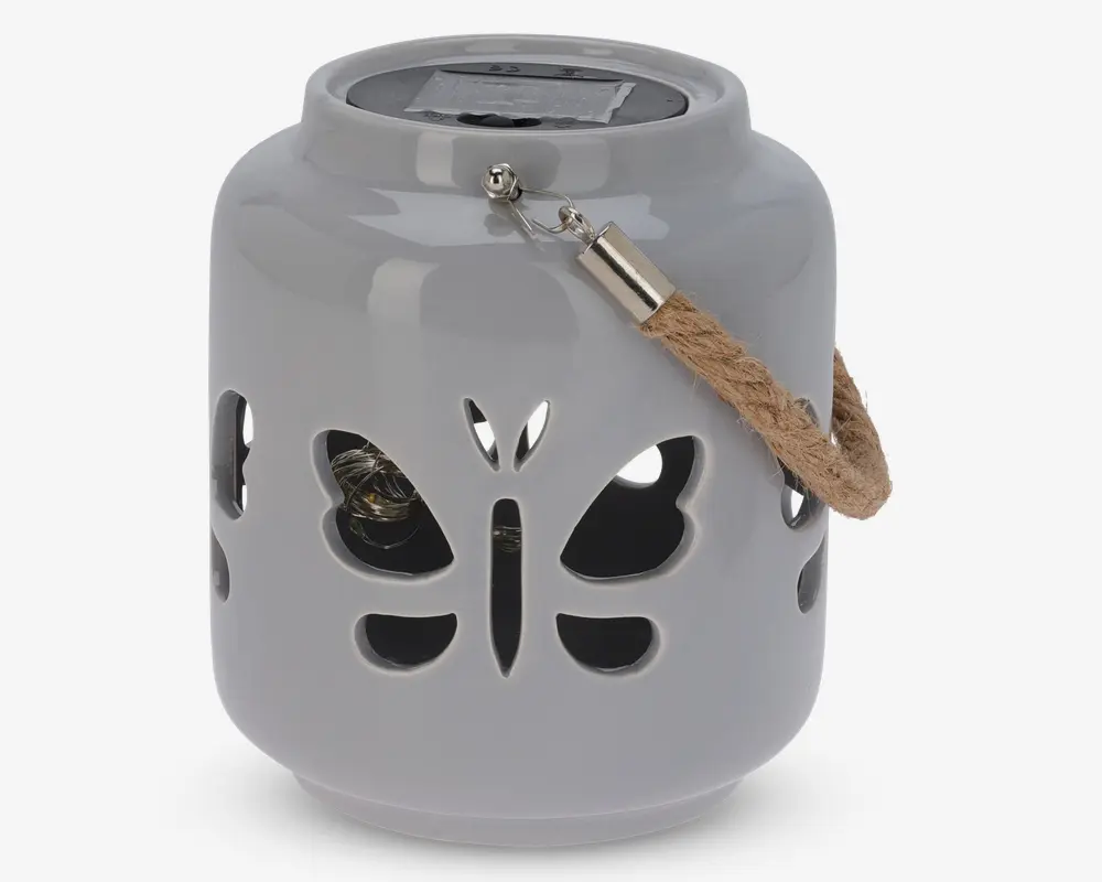 En rund, blank keramiklanterne med udskårne sommerfugleformer på siderne. Den har et robust rebhåndtag fastgjort med metalbeslag og et solpanel på toppen til opladning. Indvendigt ses en lille pære gennem udskæringerne, som er designet til at skabe dekorative lysmønstre, når lanternen er tændt.