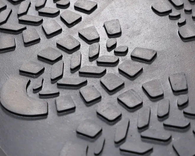 Nærbillede af en tekstureret gummioverflade med et hævet, geometrisk slidbanemønster. Mønsteret består af forskellige kantede og uregelmæssigt formede blokke, der er designet til at give greb og trækkraft. Overfladen virker slidstærk og er sandsynligvis beregnet til kraftig eller udendørs brug.
