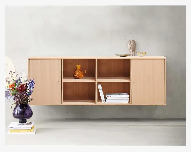 Vægmonteret skab i lyst træ med minimalistisk design, to lukkede rum med låger i hver side og fire åbne hylder i midten. På de åbne hylder står en ravfarvet glaskande med struktur, en stak bøger og et magasin. Skabet har rene linjer og en glat overflade, der kombinerer moderne og skandinavisk stil.