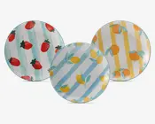 Sæt med tre runde tallerkener, hver med et forskelligt farverigt frugtmotiv. Den ene tallerken har røde jordbær med grønne blade på en baggrund af lyseblå lodrette striber. En anden tallerken viser gule citroner med grønne blade på lyseblå og hvide diagonale striber. Den tredje tallerken har orange appelsiner med grønne blade på gule og hvide lodrette striber. Alle tallerkener har en blank overflade og et legende, sommerligt udtryk.