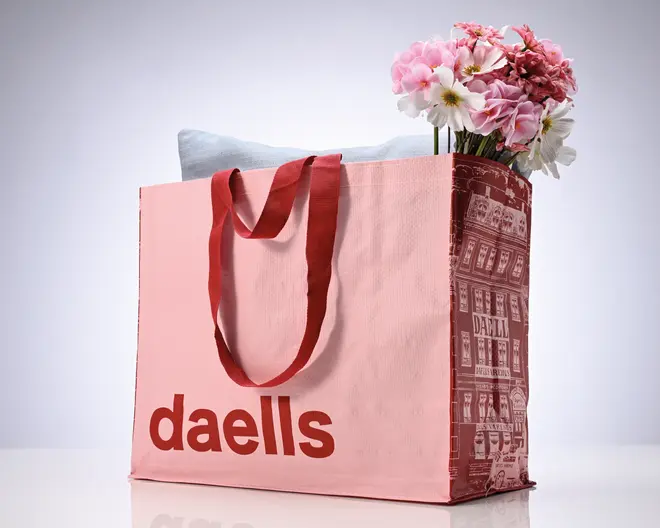 Stor lyserød totepose med lange røde båndhåndtag, en bordeauxfarvet illustreret side og teksten "daells" trykt på forsiden. Posen er vist fyldt med blomster og en lyseblå pude.