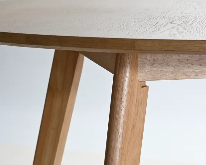 Nærbillede af en lys træbordplade med glat åretegning og skråtstillede ben i træ, hvor samlingen under bordpladen og det enkle, minimalistiske design er synligt.