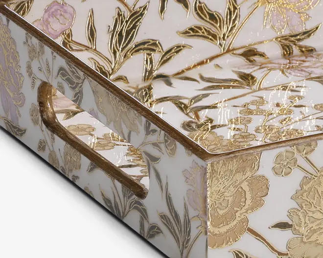 Rektangulær bakke med blank hvid overflade, dekoreret med et indviklet blomstermønster i guld og diskrete pastelfarver. Bakken har indbyggede håndtag på siderne, som også er udsmykket med det samme blomstermotiv. Gulddetaljerne er hævede, hvilket giver bakken en tekstureret og luksuriøs fremtoning.