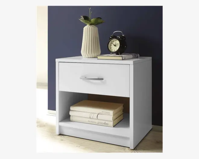 Et moderne, hvidt natbord med en enkelt skuffe, der har et stilrent sølvhåndtag. Under skuffen er der en åben hylde, hvor der ligger flere stablede bøger. Bordpladen er dekoreret med en rillet keramikvase med en grøn plante, et klassisk sort vækkeur og en lukket bog. Designet er minimalistisk og funktionelt, velegnet til moderne indretning.