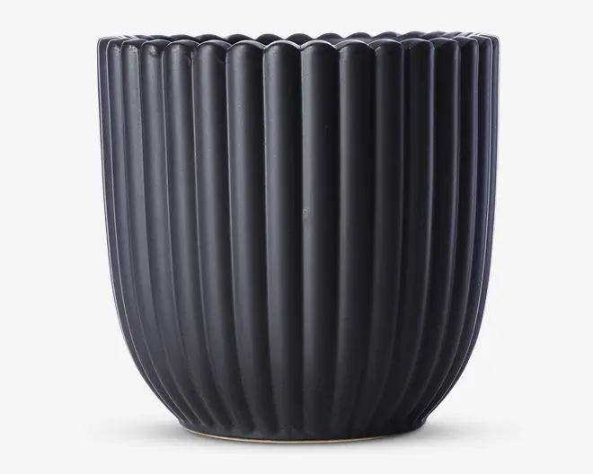 En mat sort beholder med lodrette riller og en afrundet, let tilspidset form. Kanten har en blødt bølget dekoration, som giver et elegant udtryk. Overfladen er glat, og designet er moderne og minimalistisk.