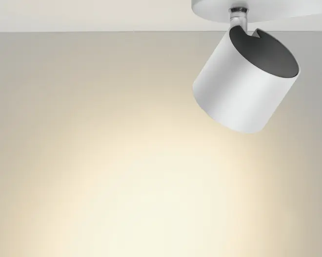 Hvid justerbar loftspot med sort inderside, monteret på en rund loftbase og vinklet, så den kaster et varmt lys på væggen.