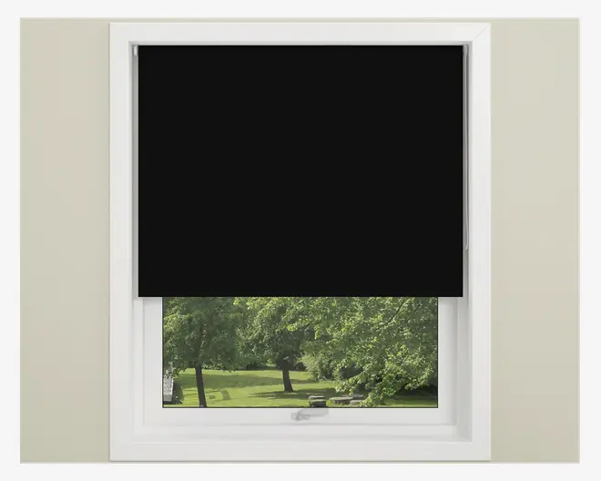 Et vindue med en sort rullegardin, der er trukket halvt ned og dækker den øverste halvdel af vinduet. Gardinet har en glat, ensfarvet sort overflade og er monteret inden for en hvid vinduesramme. Den nederste halvdel af vinduet er fri, så man kan se ud til et grønt udendørs landskab med træer og græs.