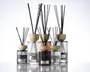 En gruppe elegante glasbeholdere med lodrette rillede strukturer, hver med et lyst trælåg og fyldt med sorte duftpinde. Beholderne varierer i størrelse og farve, herunder klare, røggrå og ravfarvede nuancer. Hver beholder har en metallabel på forsiden, hvilket giver et eksklusivt og sofistikeret udtryk.