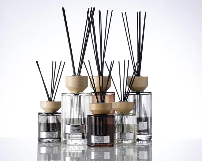 En gruppe elegante glasbeholdere med lodrette rillede strukturer, hver med et lyst trælåg og fyldt med sorte duftpinde. Beholderne varierer i størrelse og farve, herunder klare, røggrå og ravfarvede nuancer. Hver beholder har en metallabel på forsiden, hvilket giver et eksklusivt og sofistikeret udtryk.