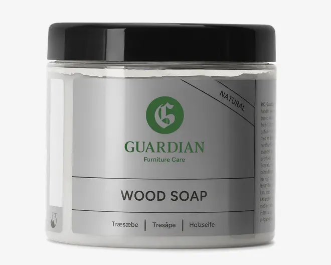En gennemsigtig plastbeholder med sort skruelåg, der indeholder en cremet, hvid sæbe. Etiketten er sølvfarvet og hvid med grøn og sort tekst og viser Guardian Furniture Care-logoet. Produktet er mærket som 'Natural' og 'Wood Soap'. Etiketten indeholder også oversættelser af 'træsæbe' på svensk og tysk. Beholderen har et moderne, minimalistisk design med rene linjer og et professionelt udtryk.