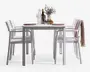 Et moderne udendørs spisesæt bestående af et rektangulært hvidt bord med fire matchende hvide stole. Stolene har lamel-sæder og rygge samt armlæn for ekstra komfort. Bordet er dækket op til to personer med lyserøde tallerkener, vinglas og bestik. Midt på bordet står en klar glaskaraffel, en vinflaske, to ekstra glas, en lille vase med blomster og en skål med frugt. En af stolene har et lyserødt, tekstureret tæppe hængende over ryggen, hvilket giver et hyggeligt præg.