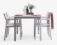 Et moderne udendørs spisesæt bestående af et rektangulært hvidt bord med fire matchende hvide stole. Stolene har lamel-sæder og rygge samt armlæn for ekstra komfort. Bordet er dækket op til to personer med lyserøde tallerkener, vinglas og bestik. Midt på bordet står en klar glaskaraffel, en vinflaske, to ekstra glas, en lille vase med blomster og en skål med frugt. En af stolene har et lyserødt, tekstureret tæppe hængende over ryggen, hvilket giver et hyggeligt præg.