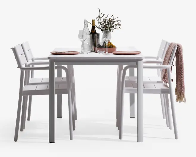 Et moderne udendørs spisesæt bestående af et rektangulært hvidt bord med fire matchende hvide stole. Stolene har lamel-sæder og rygge samt armlæn for ekstra komfort. Bordet er dækket op til to personer med lyserøde tallerkener, vinglas og bestik. Midt på bordet står en klar glaskaraffel, en vinflaske, to ekstra glas, en lille vase med blomster og en skål med frugt. En af stolene har et lyserødt, tekstureret tæppe hængende over ryggen, hvilket giver et hyggeligt præg.
