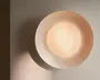 Rund vægmonteret lampe med varm beige yderringsdetalje og en blødt lysende midte, der kaster en mild skygge på væggen.