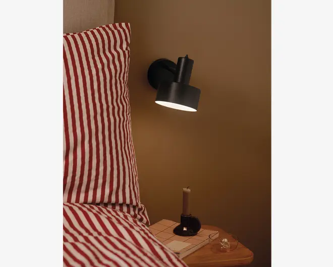 En moderne sort væglampe med cylindrisk skærm, monteret over et sengebord. Lampen udsender et varmt, fokuseret lys nedad. Sengebordet har en lille sort lysestage med et beige stearinlys, et par briller og en bog med ternet omslag. Sengen ved siden af bordet har rødt og hvidt stribet sengetøj.