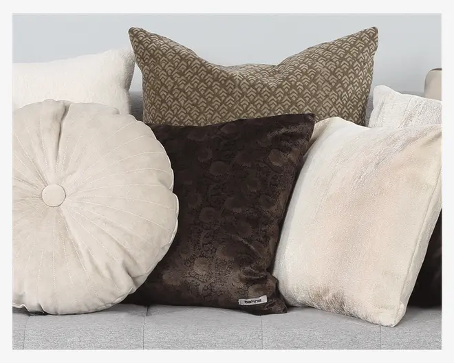 En samling dekorative puder i forskellige former, størrelser og teksturer. Arrangementet inkluderer en rund, cremefarvet pude med syede radiære linjer, en firkantet pude med mørkebrun blomsterstruktur, en stor firkantet pude med geometrisk beige og brunt mønster samt to firkantede puder i bløde, lyse beige og offwhite stoffer. Pudernes materialer varierer mellem velour, tekstur og mønster, hvilket skaber et hyggeligt og stilfuldt udtryk.