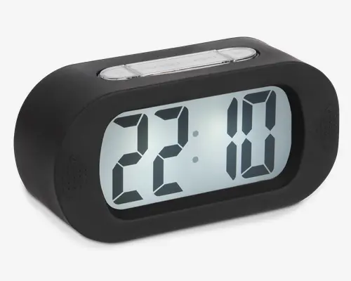 Rektangulært digitalur med afrundede kanter, der har et stort, letlæseligt LCD-display, som viser tiden i 24-timers format. Uret har et sort kabinet og en markant sølvfarvet knap på toppen, sandsynligvis til snooze- eller lysfunktion. Displayet er baggrundsbelyst for synlighed i svagt lys.