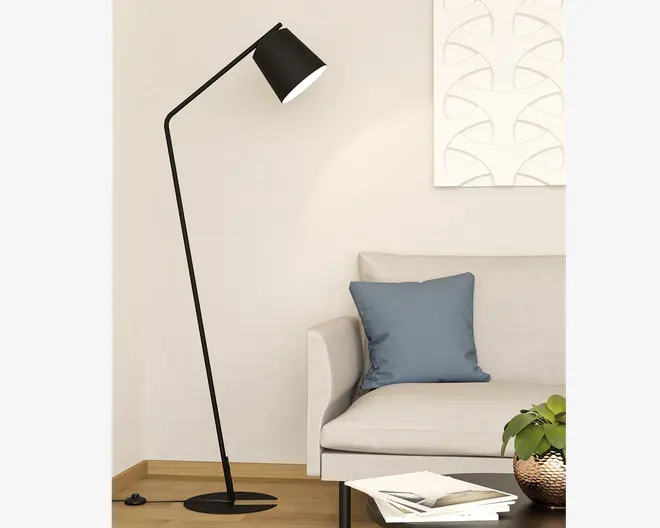Høj, moderne gulvlampe med mat sort finish og et vinklet, minimalistisk design. Lampen har en slank, lige stang, der bøjer nær toppen og bærer en konisk lampeskærm med hvid inderside for bedre lysrefleksion. Lampen står på en rund, flad fod for stabilitet og er placeret ved siden af en lysefarvet sofa i en moderne stue.