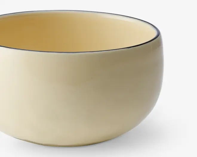 En rund, glat keramikskål med blank cremefarvet overflade både indvendigt og udvendigt. Skålens kant er markeret med en tynd, mørk streg, som giver et diskret dekorativt præg. Skålen har et enkelt, minimalistisk design uden hank eller mønster.