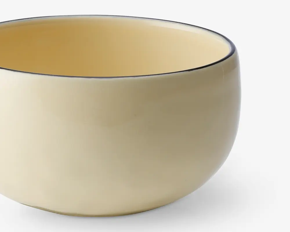 En rund, glat keramikskål med blank cremefarvet overflade både indvendigt og udvendigt. Skålens kant er markeret med en tynd, mørk streg, som giver et diskret dekorativt præg. Skålen har et enkelt, minimalistisk design uden hank eller mønster.