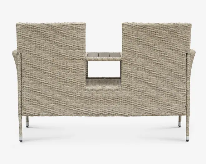 Bagside af en udendørs bænk med to sæder, lavet af lysebeige flettet rattan. Bænken har et unikt design med et rektangulært udskåret område i midten af ryglænet, som afslører et lille indbygget bord eller hylde mellem de to siddepladser. Benene er slanke og lige, og matcher bænkens flettede struktur.