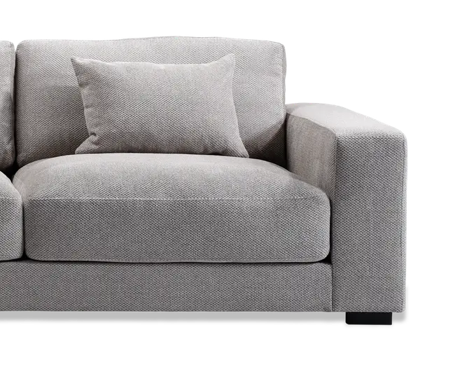 Nærbillede af en moderne sofa med en kraftig, firkantet armlæn og bløde sæde- og ryghynder. Betrækket er i et lysegråt, tekstureret stof, og der er en matchende pyntepude placeret mod ryglænet. Designet er minimalistisk og moderne med rene linjer og et robust udtryk.