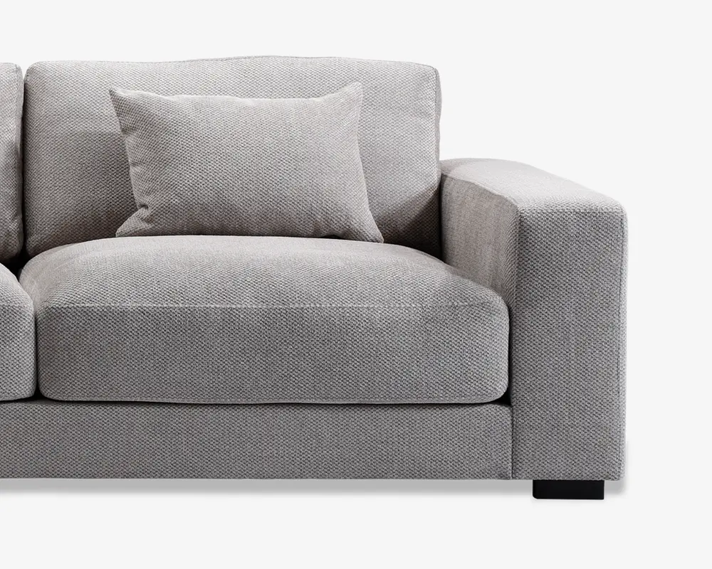 Nærbillede af en moderne sofa med en kraftig, firkantet armlæn og bløde sæde- og ryghynder. Betrækket er i et lysegråt, tekstureret stof, og der er en matchende pyntepude placeret mod ryglænet. Designet er minimalistisk og moderne med rene linjer og et robust udtryk.