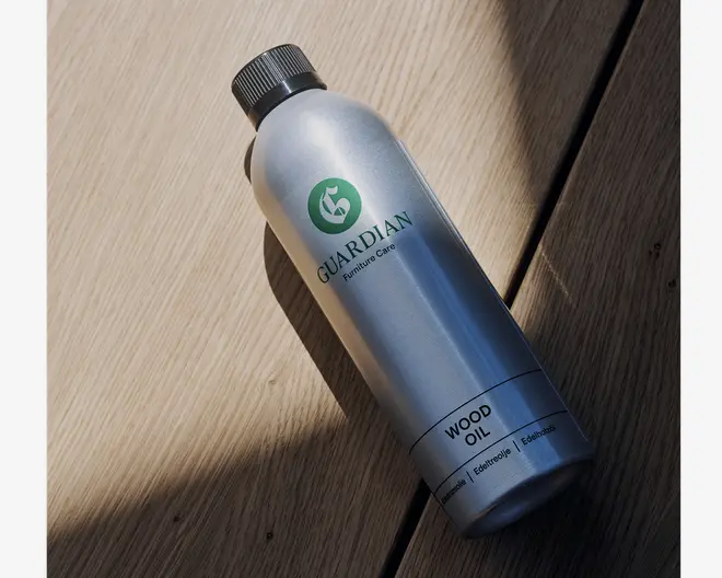 En slank, cylindrisk sølvfarvet metalflaske med sort skruelåg. Flasken har et grønt, rundt logo med et hvidt 'G' og teksten 'GUARDIAN Furniture Care' trykt nedenunder. Nederst på flasken står der 'WOOD OIL' med store bogstaver, samt yderligere tekst, der angiver egnethed til forskellige træsorter. Flasken ligger på en lys træoverflade, og naturligt lys kaster bløde skygger.